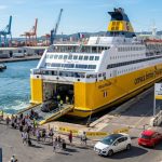 Un traghetto Corsica Ferries giallo attraccato al porto di Sète, in Francia, con passeggeri e veicoli che si imbarcano. (Immagine generata con IA)