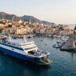 Un traghetto si avvicina o lascia un porto isolano italiano, con edifici tipici mediterranei e piccole imbarcazioni. (Immagine generata con IA)