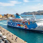 Un traghetto moderno di Moby, Tirrenia o Toremar salpa da un porto turistico animato, simboleggiando la ripartenza della stagione 2027. (Immagine generata con IA)