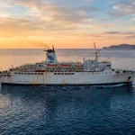 Un grande traghetto passeggeri simile al GNV Azzurra naviga al tramonto, con una luce dorata che ne evidenzia la struttura. (Immagine generata con IA)
